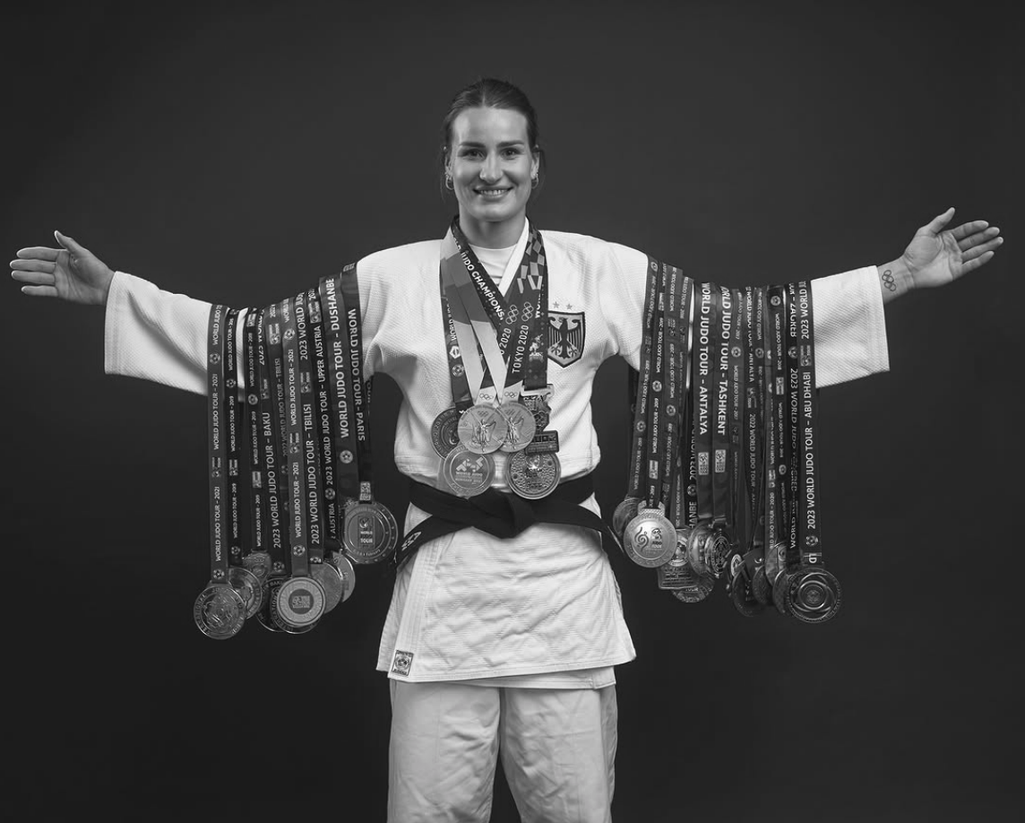 Judoka in Sportkleidung präsentiert zahlreiche Medaillen vor dunklem Hintergrund.